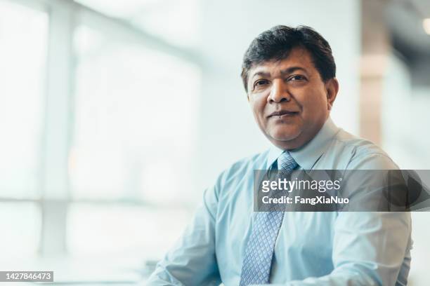 portrait of asian indian mature businessman sitting in bright office in businesswear - indian old man stockfoto's en -beelden