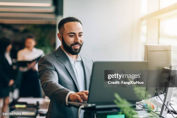 multirassischer portugiesischer jamaikanischer mittelerwachsener geschäftsmann mit bart, der lächelnd am schreibtisch mit laptop steht und daten im hellen geschäftsbüro im anzug überprüft - reading laptop pleased stock-fotos und bilder