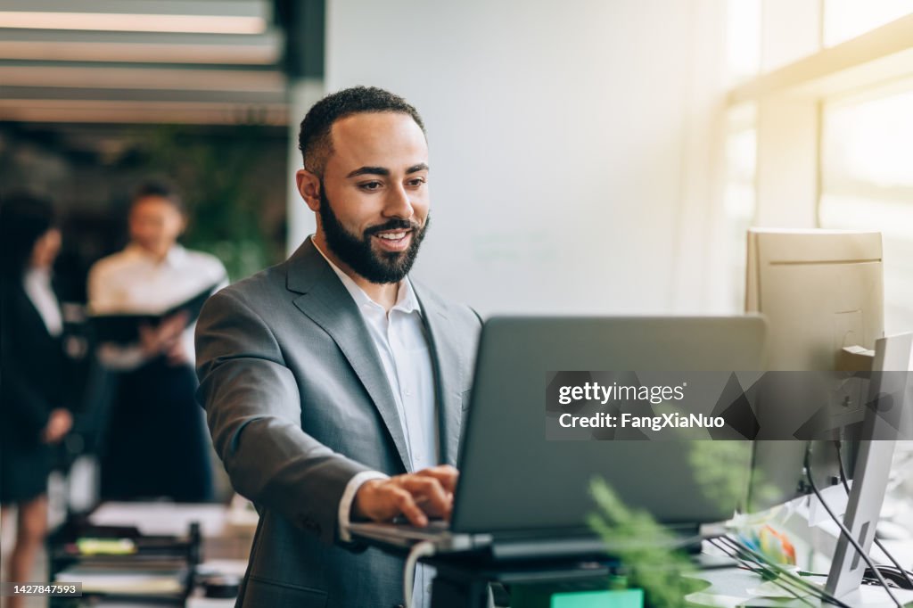Multiracial Portuguese Jamaican mid adult businessman with beard standing smiling at desk with laptop reviewing data in bright business office wearing suit