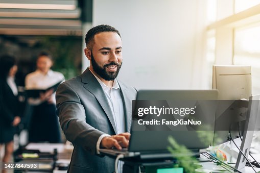 Multiracial Portuguese Jamaican mid adult businessman with beard standing smiling at desk with laptop reviewing data in bright business office wearing suit Multiracial Portuguese Jamaican mid adult businessman with beard standing smiling at desk with laptop reviewing data in bright business office wearing suit