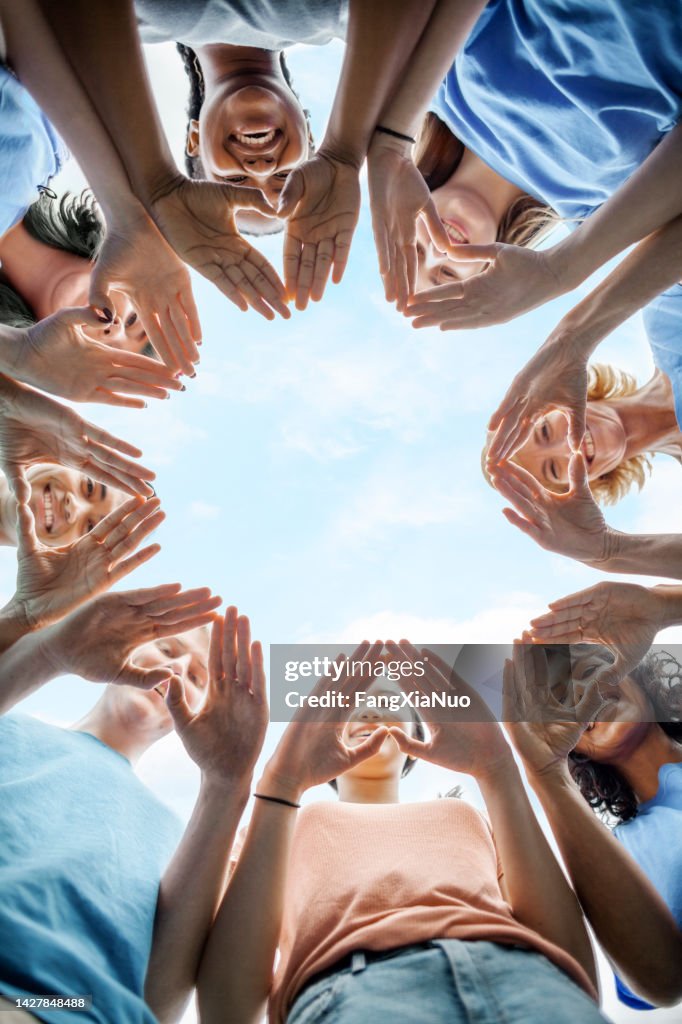 Multiracial group of diverse people stand in circle as community volunteers to show support and commitment to teamwork success togetherness making hand gesture in concept symbol sign of heart shape outdoors with sky