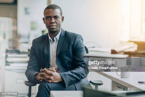 portrait of black mid adult businessman student sitting serious with hands clasped in bright business office wearing suit - black suit 個照片及圖片檔