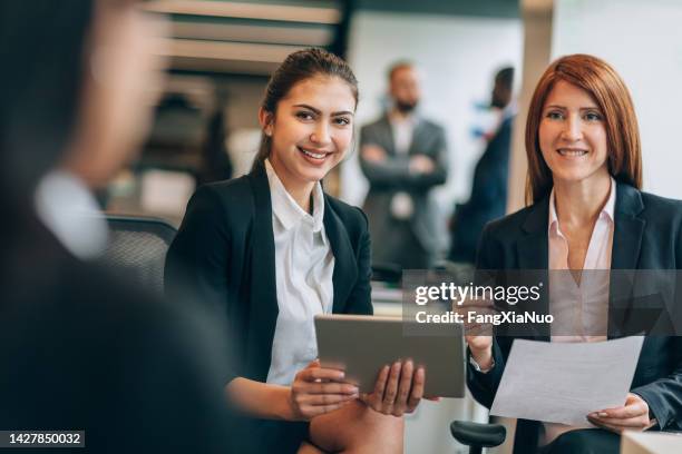 pair of mid adult businesswomen talking discussing smiling holding tablet computer with colleague in business meeting in office wearing suits businesswear - hr suit stockfoto's en -beelden