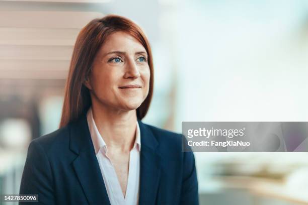 portrait of caucasian redhead mature businesswoman student smiling contented emotion satisfaction aspiration looking away forward in bright business office wearing suit businesswear - three quarter front view 個照片及圖片檔