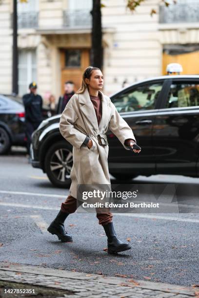 Guest wears a black shiny varnished leather headband, a dark brown sweater, a beige belted long coat, dark brown sport pants, black shiny leather...