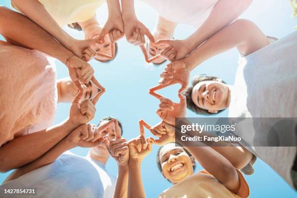 multiracial group of diverse young people students stand in circle as community volunteers to show support and commitment to teamwork success togetherness making hand gesture in concept symbol sign of heart shape outdoors with sky - team t shirt imagens e fotografias de stock