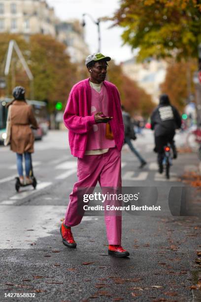 Guest wears white mesh and green bicolored cap, gold earrings, a pale yellow t-shirt, a pink wool V-neck with pale pink circle print pattern...