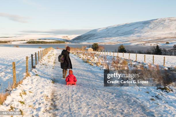 mother wearing a winter jacket and a woolly hat pulls a sledge with her daughter in it across the ice and snow in the pentland hills regional park in edinburgh, scotland, uk - edinburgh scotland photos et images de collection