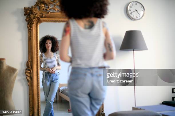 young tattooed woman with curly hair looking at her reflection in full length mirror at home - woman full length mirror stock pictures, royalty-free photos & images