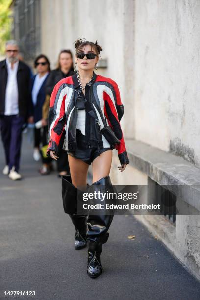 Guest wears black sunglasses, silver earrings, a black / red / white shiny leather oversized biker jacket, a black shiny leather Triangular crossbody...