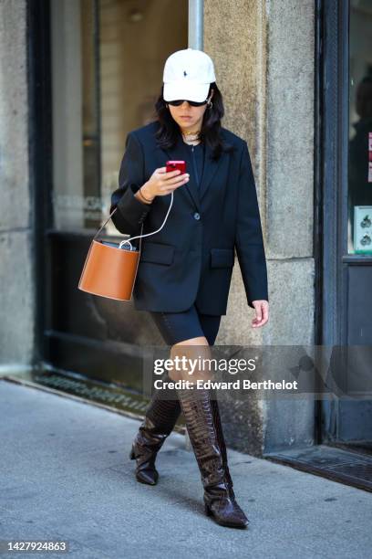 Guest wears a white with black Ralph Lauren embroidered pattern cap, gold earrings, a black blazer jacket, a black zipper t-shirt, black cyclist...