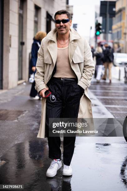 Alex Badia is seen wearing a beige fishnet tank-top, a beige long trench coat, a black and white striped suit pants outside the Salvatore Ferragamo...