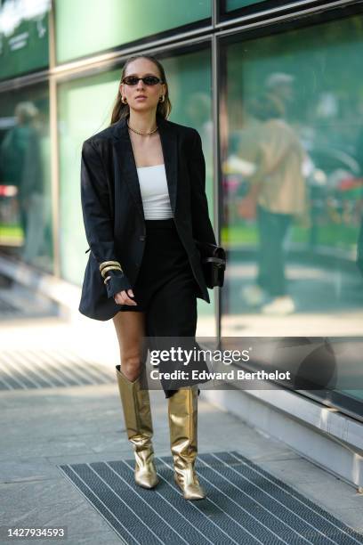 Guest wears black sunglasses, a gold necklace, a white shoulder-off top, a black blazer jacket, a gold large bracelet, a black asymmetric skirt, gold...