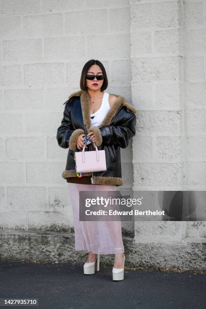 Guest wears black sunglasses, a white tank-top from Prada, a black shiny leather with embroidered brown fur interior oversized aviator coat, a pale...