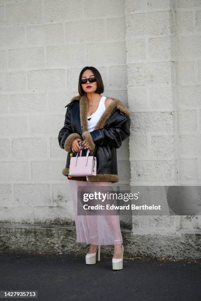 Guest wears black sunglasses, a white tank-top from Prada, a black shiny leather with embroidered brown fur interior oversized aviator coat, a pale...