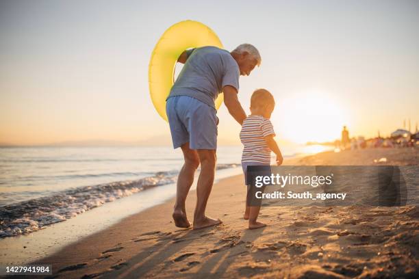 grand-père marchant sur la plage avec petit-fils et anneau gonflable au coucher du soleil - grand parent photos et images de collection