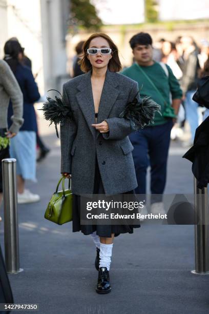 Guest wears white sunglasses, a black bra underwear, a dark gray oversized blazer jacket with embroidered feathers sleeves, a dark gray knees pleated...