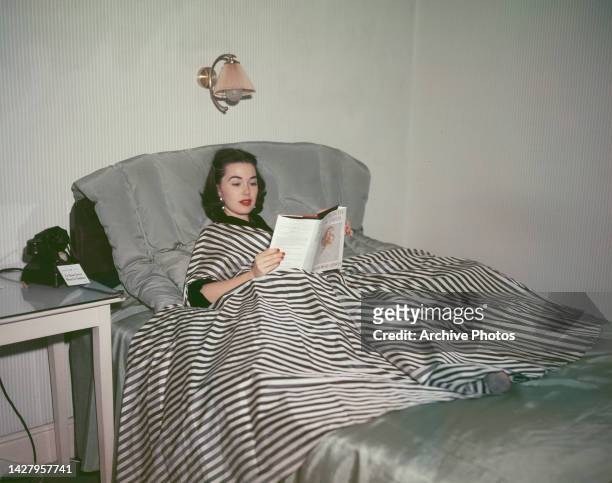 American actress Barbara Rush, wearing a black-and-white striped outfit, lying on a bed reading a copy of 'A Month in Paris' by Mrs Robert Henrey ,...