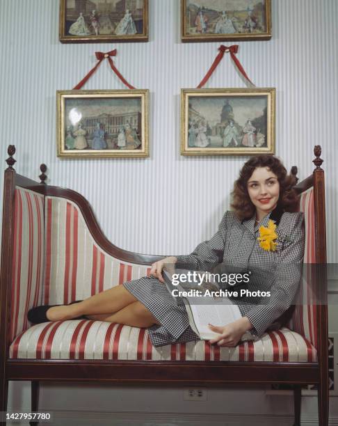 American actress Jennifer Jones wearing a black-and-white checked outfit with daffodil attached to the front, as holds an open book in her lap while...