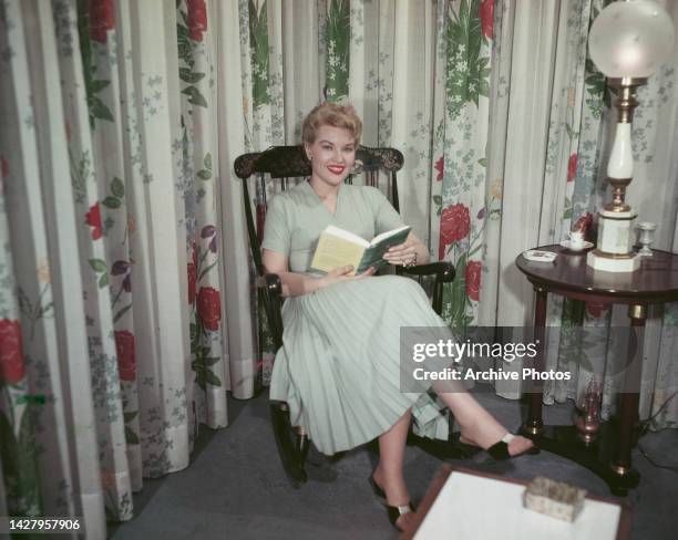 American singer and actress Patti Page , wearing a pale green dress, sits reading a book in a rocking chair in front of curtains with a floral...