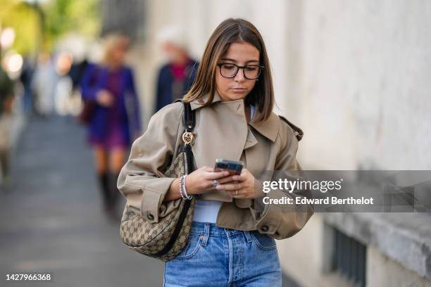 Guest wears black glasses, a beige cropped jacket, a beige and brown GG monogram print pattern shoulder bag from Gucci, bracelets, blue faded denim...