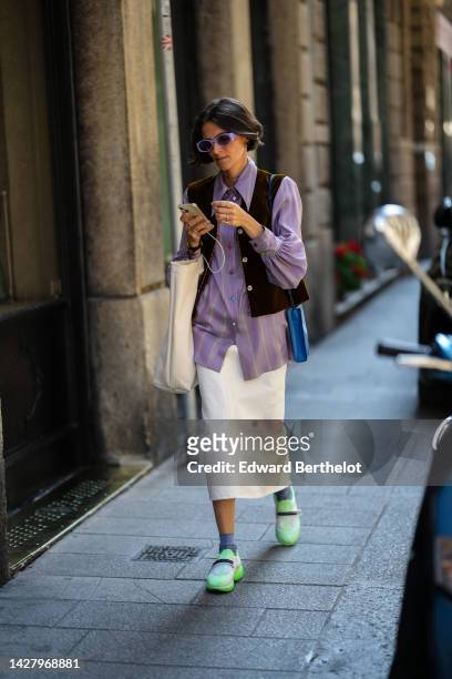 Guest wears purple sunglasses, gold necklaces, a purple striped print pattern shirt, a black sleeveless gilet, silver and gold rings, a blue shiny...