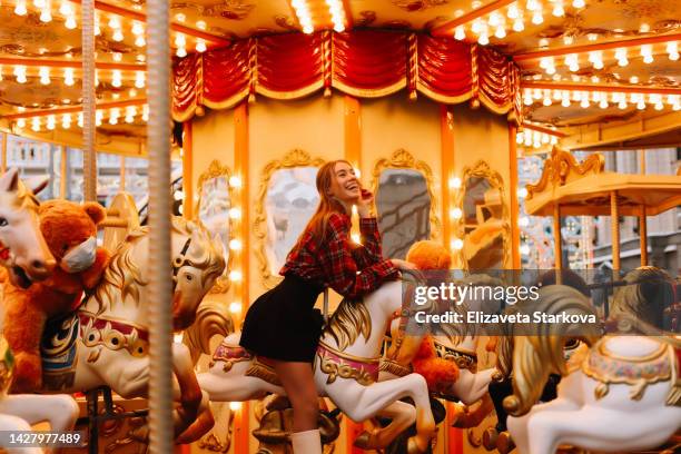 a beautiful smiling young woman rides a merry-go-round with horses on a christmas holiday on a decorated street celebrating the new year in winter outdoor - carousel horse stock pictures, royalty-free photos & images