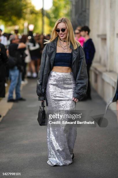 Guest wears black sunglasses, silver large chain necklaces, a navy blue denim shoulder-off / cropped top, a black shiny leather oversized bomber...