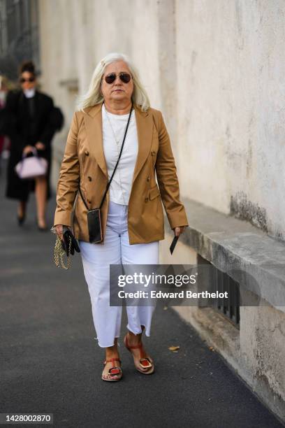 Guest wears black Aviator sunglasses, a gold long chain necklace, a white t-shirt, a brown blazer jacket, white denim large pants, a black shiny...