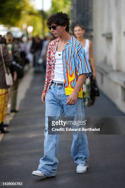Guest wears black sunglasses, a white tank-top from Prada, a blue and red striped print pattern and half red and white fire print pattern shirt, a...