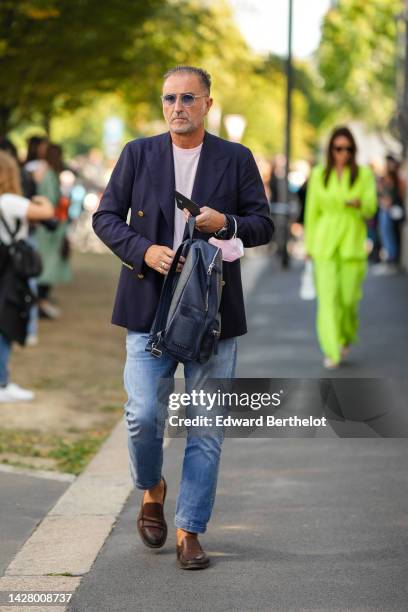 Guest wears blue sunglasses, a white t-shirt, a navy blue blazer jacket with gold buttons, a navy blue shiny leather back pack bag, blue faded denim...