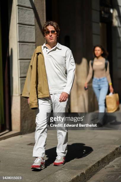 Guest wears brown sunglasses, a white long sleeves polo shirt, a pale khaki zipper jacket, white large pants, pale gray and red suede sneakers from...