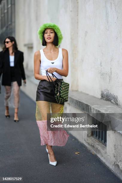 Molly Chiang wears a green fluffy bob hat, a white tank-top from Prada, a multi materials black shiny leather / yellow tulle / pink tulle long skirt...