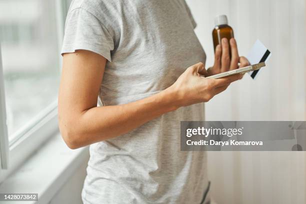 a woman in a gray home t-shirt stands and holds a phone and bottle with liquid medicine in one hand and a credit card in the other. ordering medicine on the internet, searching for information, online ordering with delivery. - prescription home delivery stock pictures, royalty-free photos & images