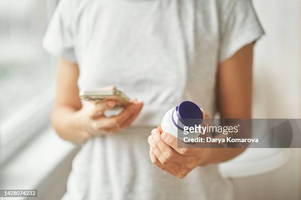 a woman in a gray home t-shirt stands and holds a phone in one hand and tablets in the other. ordering pills on the internet, searching for medicines and information, online ordering home with delivery. - dr t and the women stock-fotos und bilder