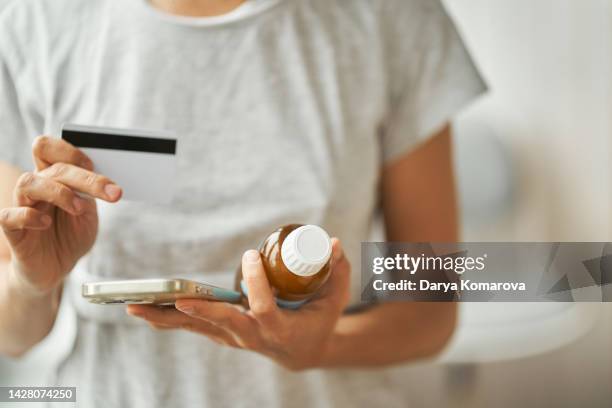 a woman in a gray home t-shirt stands and holds a phone and bottle with liquid medicine in one hand and a credit card in the other. ordering medicine on the internet, searching for information, online ordering with delivery. - dr t and the women stock-fotos und bilder