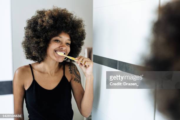 multiracial woman brushing teeth in bathroom in front of the mirror - teeth cleaning stock pictures, royalty-free photos & images