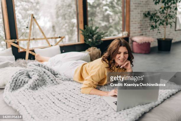 young beautiful asian woman with dark long hair in cozy white knitted sweater using laptop on bed in room with christmas tree. portrait of carefree happy teenager girl student in house in christmas - weihnachten laptop stock-fotos und bilder