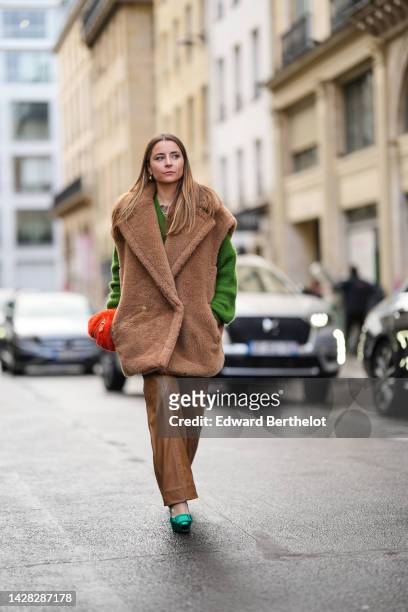Julia Comil wears gold earrings, a green V-neck wool pullover, a brown oversized fluffy sleeveless coat, brown shiny leather flared pants, a neon...