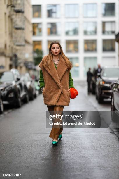 Julia Comil wears gold earrings, a green V-neck wool pullover, a brown oversized fluffy sleeveless coat, brown shiny leather flared pants, a neon...