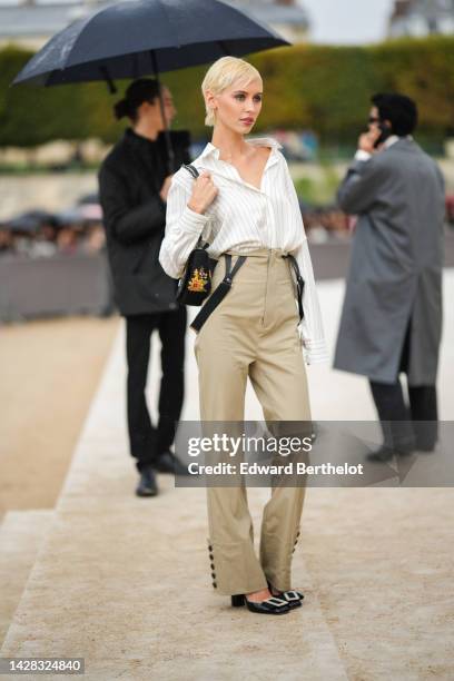 Iris Law wears a gold chain pendant earring, a whit latte with small striped print pattern shirt, a black fabric with embroidered orange / green...