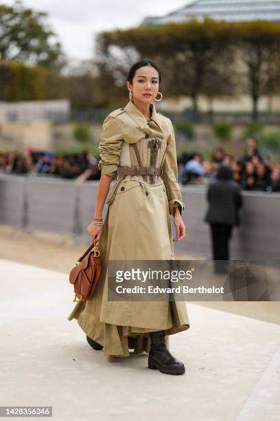 Yoyo Cao wears gold earrings, a beige long trench coat from Dior, a beige and brown laces corset, diamonds bracelets, a brown camel shiny leather...