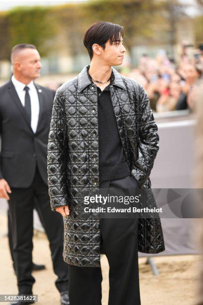 Cha Eun-woo wears a black and silver chain necklace, a black polo shirt, a black shiny varnished / embossed iconic Dior cannage coat from Dior, black...