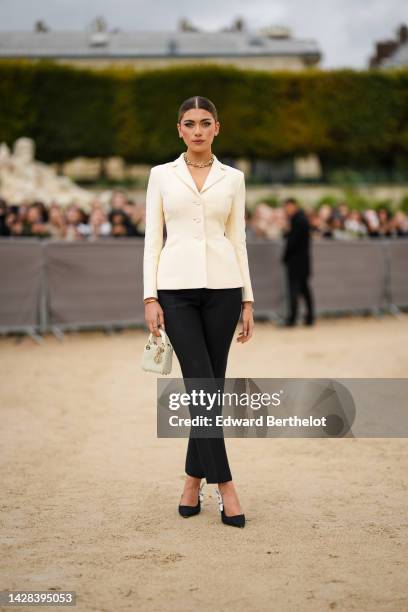 Amelie Zilber wears a gold with embroidered diamonds necklace, a white latte blazer jacket, black suit pants, a gold bracelet, a white pearls micro...