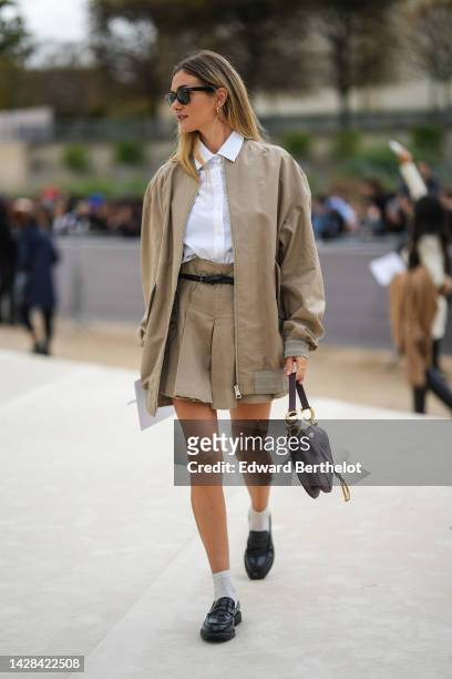 Guest wears black sunglasses from Ray Ban, gold earrings, a white shirt, a beige jacket from Dior, a matching beige pleated / accordion short skirt...