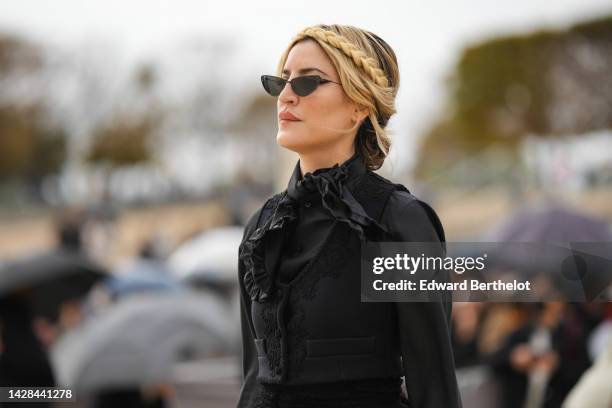 Guest wears black cat eyes sunglasses, a black knot neck / silk shirt, a black embroidered pattern sleeveless / cropped jacket, a black embroidered /...