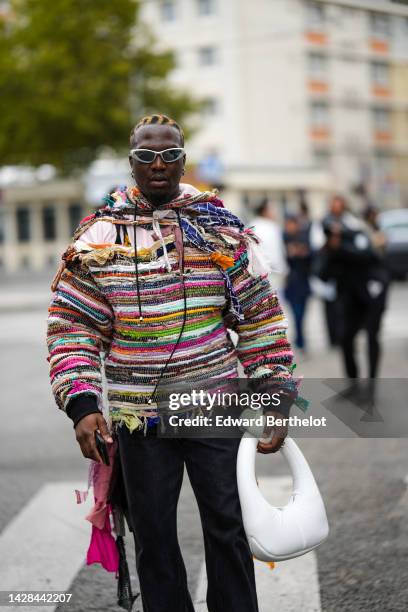 Guest wears silver sunglasses, a multicolored embroidered striped pattern ripped pullover, black denim pants, a white puffy leather handbag, outside...