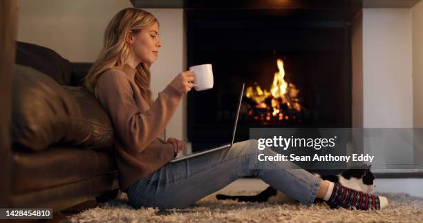 happy woman, fireplace and laptop while browsing the internet, watching movie or writing article with coffee for a blog at home. young female streaming online film or series subscription drinking tea - cosy fire stock pictures, royalty-free photos & images