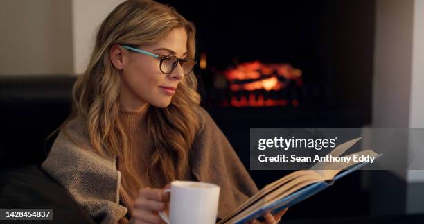 woman reading book with coffee by fireplace in living room for knowledge, leisure or studying. reader or bookworm with glasses, drinking tea and enjoying literature fiction story at night in her home - eyeglasses winter stock pictures, royalty-free photos & images