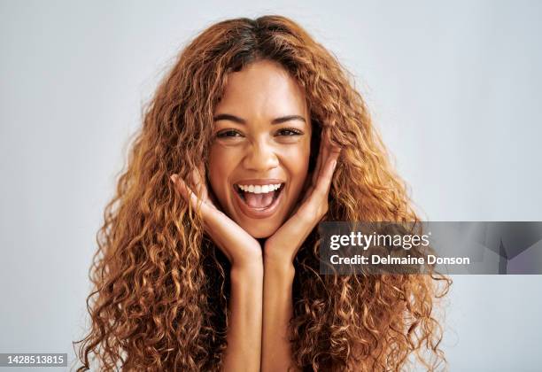 modèle aux cheveux bouclés, a la tête dans les mains tout en souriant et excité sur fond de studio gris. une jeune femme aux cheveux naturels a l’expression du bonheur et du plaisir sur fond ou mur gris - portrait of beautiful brunette woman smile photos et images de collection